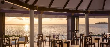 An outdoor dining area with tables and chairs, a ceiling fan, and a view of the sea at sunset.