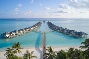 Un complejo turístico con bungalows sobre el agua, conectados por un muelle, una playa y palmeras.