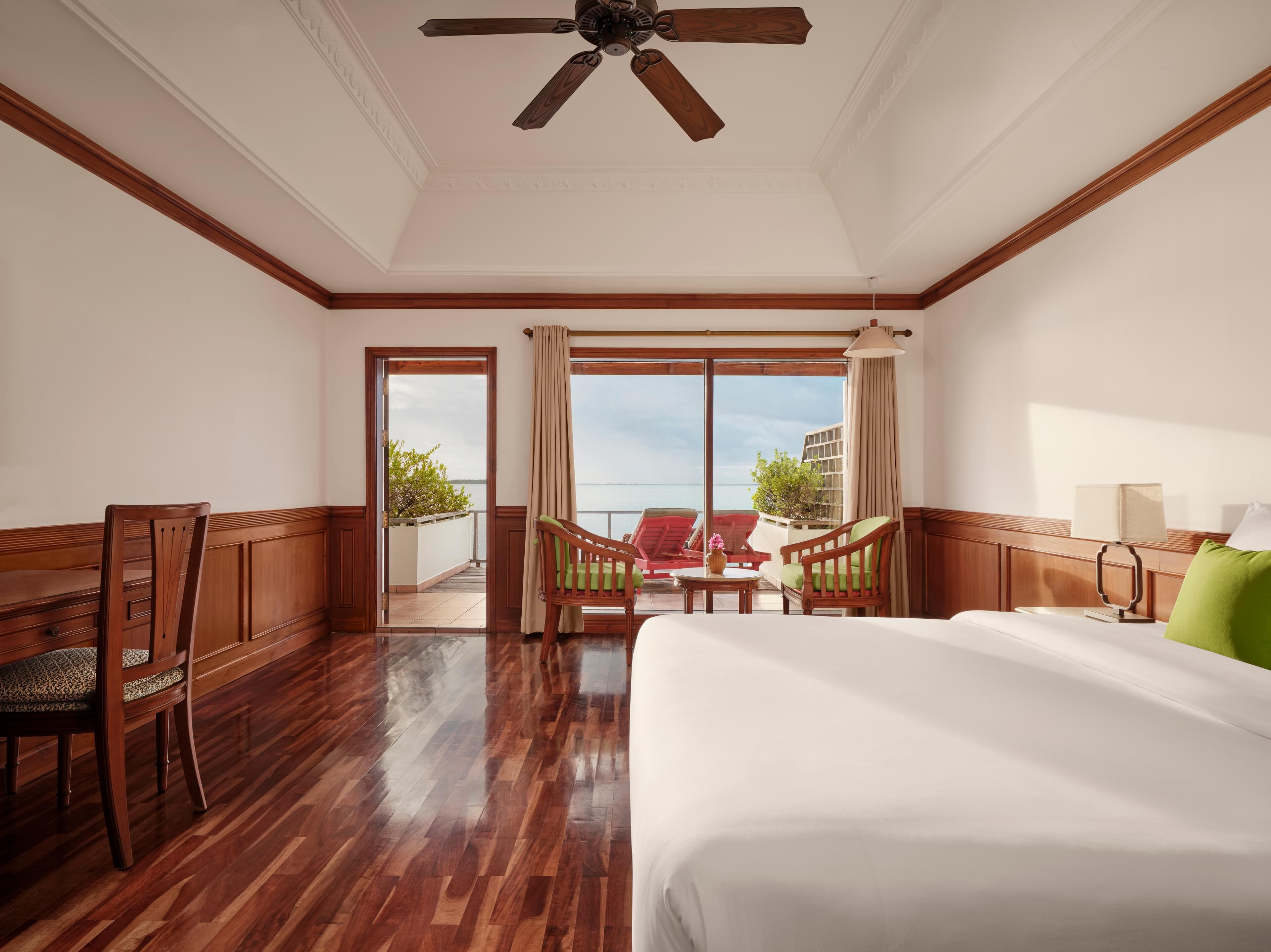 A hotel room with a large bed, wooden furniture, a ceiling fan, and a balcony with a view of the sea.