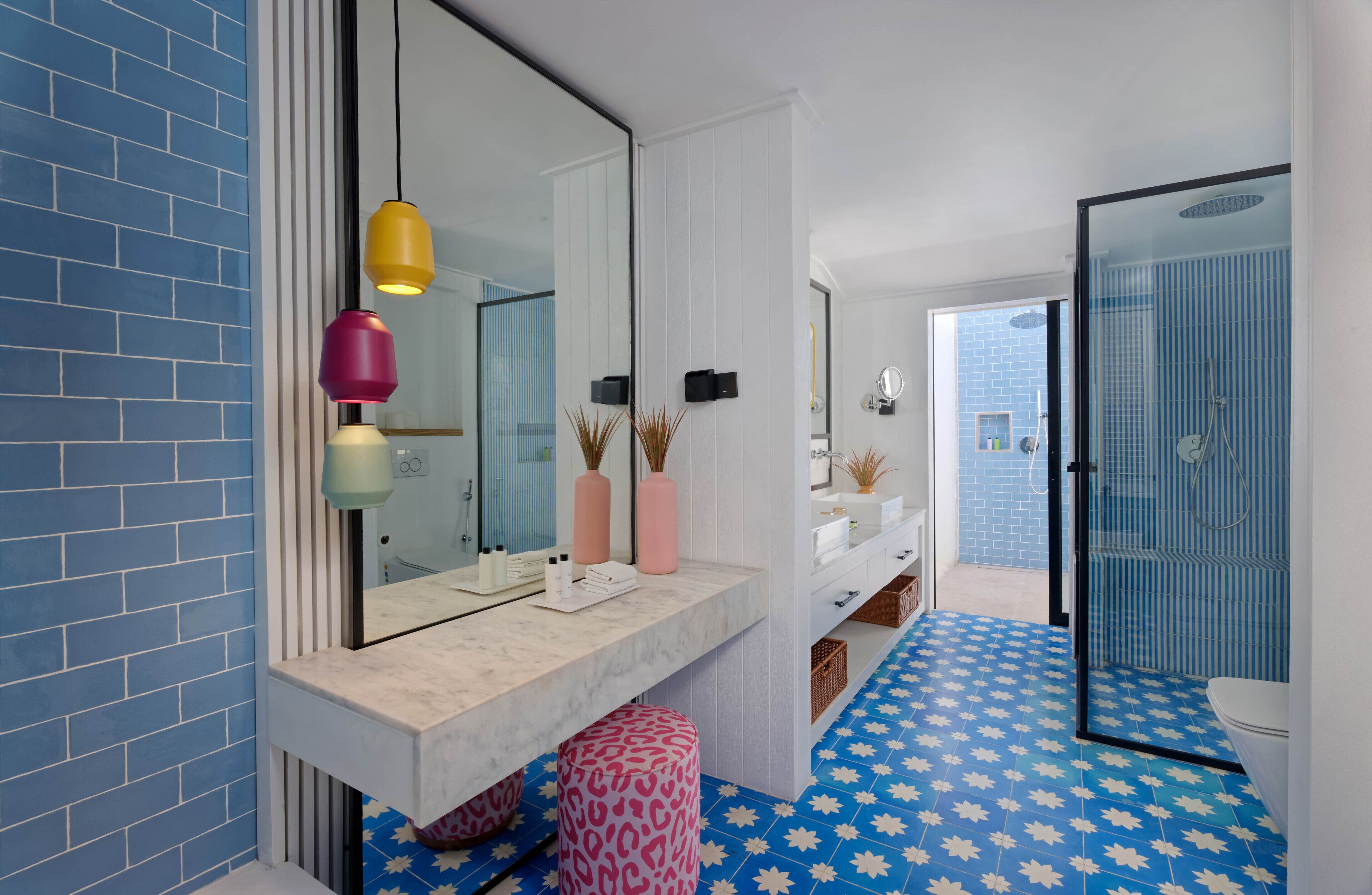 Un baño moderno con piso de baldosas azules y blancas, un espejo grande y un tocador blanco con lámparas colgantes rosadas y amarillas.