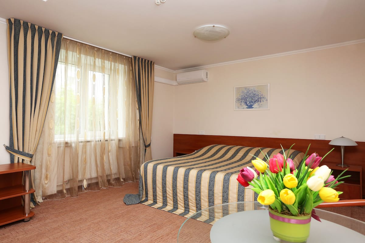 Una habitación de hotel con una cama, un escritorio, una silla, una mesa, un jarrón de flores y un cuadro en la pared.