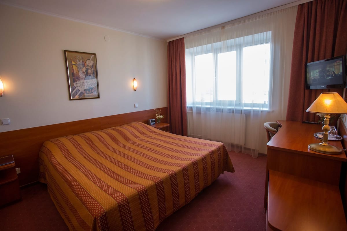 Habitación de hotel con una cama grande, un escritorio con lámpara, televisor y ventana con cortinas.