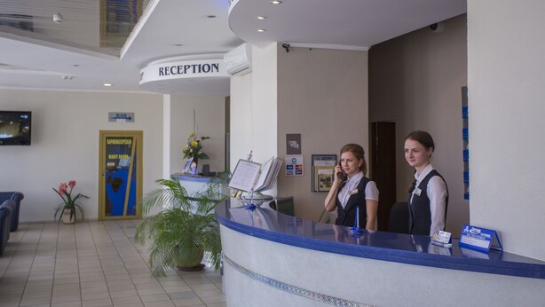 A reception desk with a blue counter, a reception sign, a digital screen, and a potted plant. - Sputnik (Minsk)