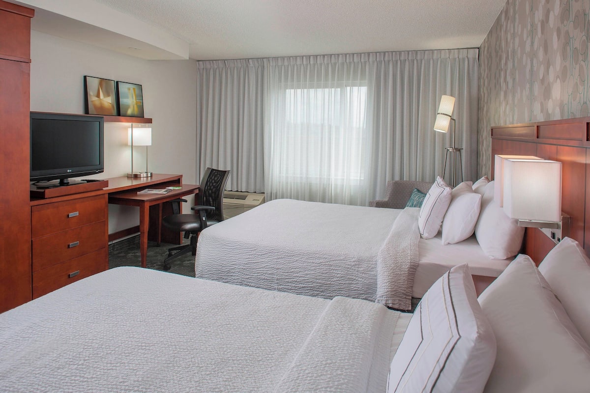 Une chambre d’hôtel avec deux lits, un bureau avec une télévision, une chaise, une lampe et une fenêtre avec des rideaux.