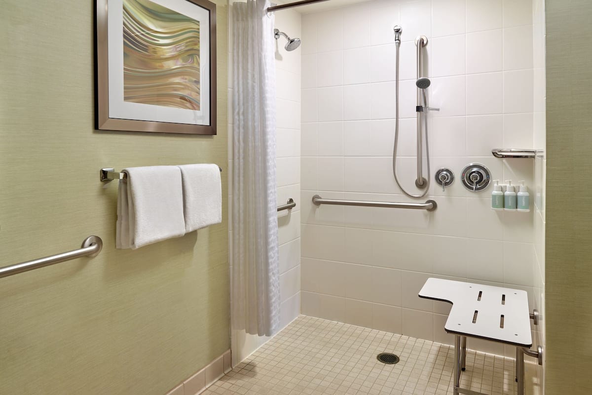 Une douche accessible aux personnes en fauteuil roulant, équipée de barres d’appui, d’un petit lavabo et d’un porte-serviettes.
