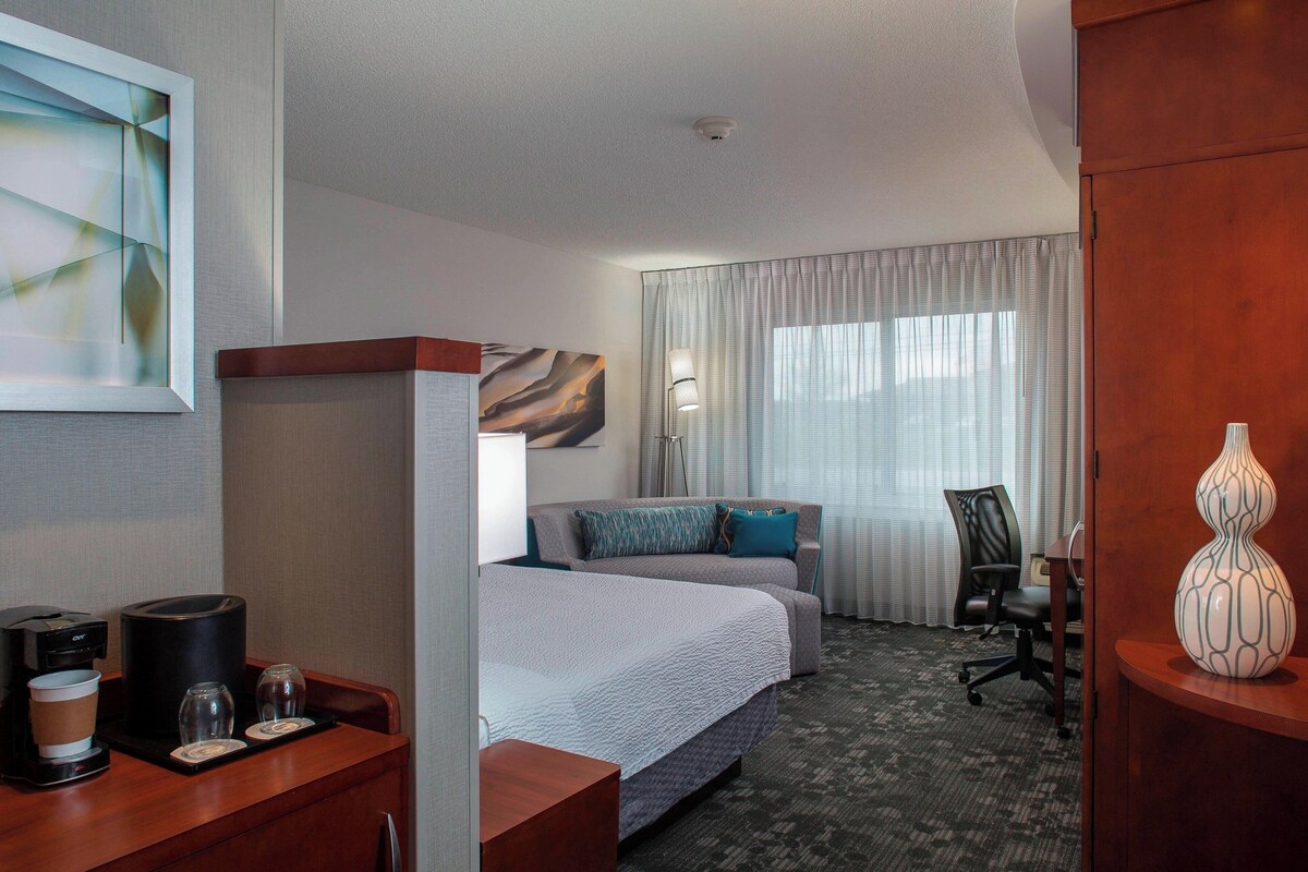A hotel room with a bed, a desk with a coffee maker, a lamp, and a chair.