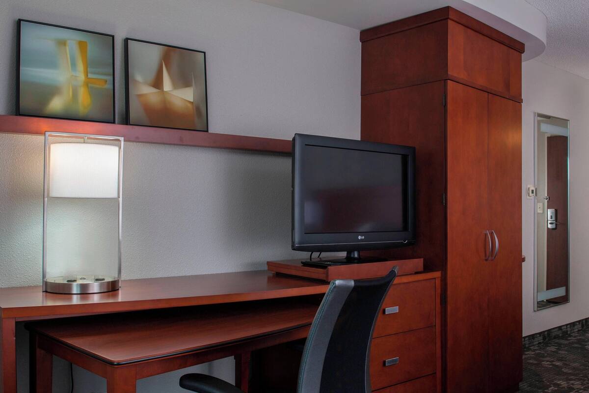 Une chambre d’hôtel avec un bureau en bois, un téléviseur à écran plat, une chaise, un placard et une lampe fixée au mur.