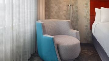 Habitación de hotel con un sillón gris, cama con ropa de cama blanca, una lámpara de pie y papel tapiz estampado.