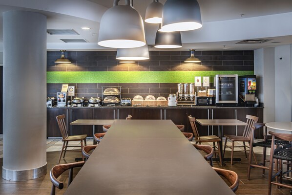 A modern restaurant with a long communal table, multiple chairs, and a counter with food and beverage dispensers.