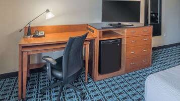 A hotel room with a wooden desk, a black office chair, a small black refrigerator, a flat-screen TV, and a bed with a white comforter.