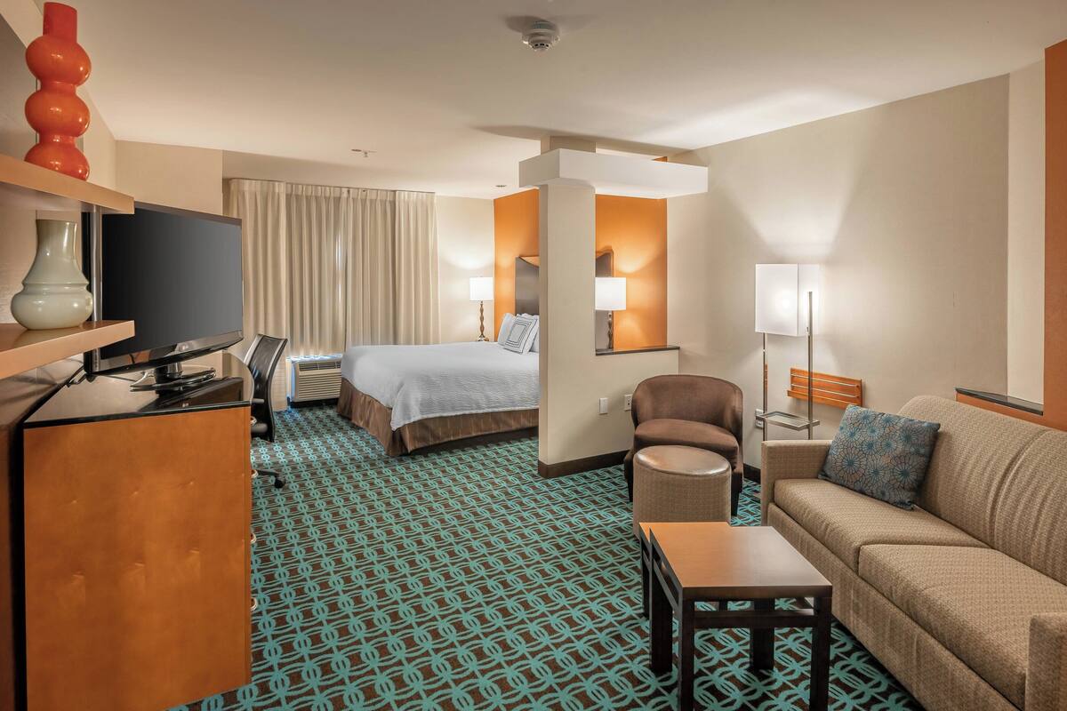 A hotel room with a bed, a sofa, a desk, a TV, and a patterned carpet.