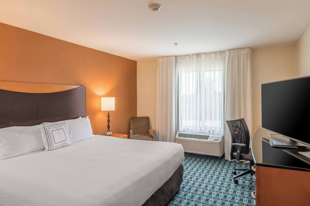 A hotel room with a large bed, a desk with a flat-screen TV, a chair, a lamp, and a window with curtains.