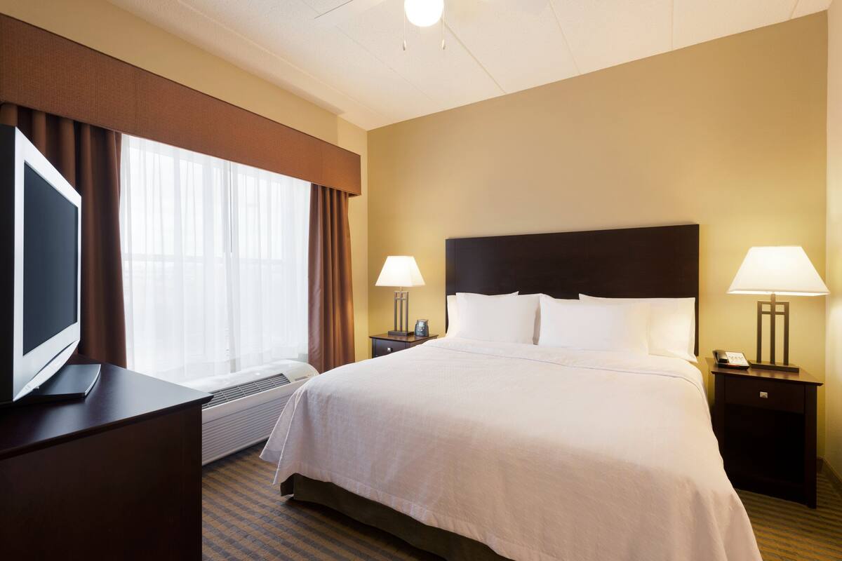 A hotel room with a large bed, two bedside tables with lamps, a flat-screen TV on a desk, and a large window with curtains.