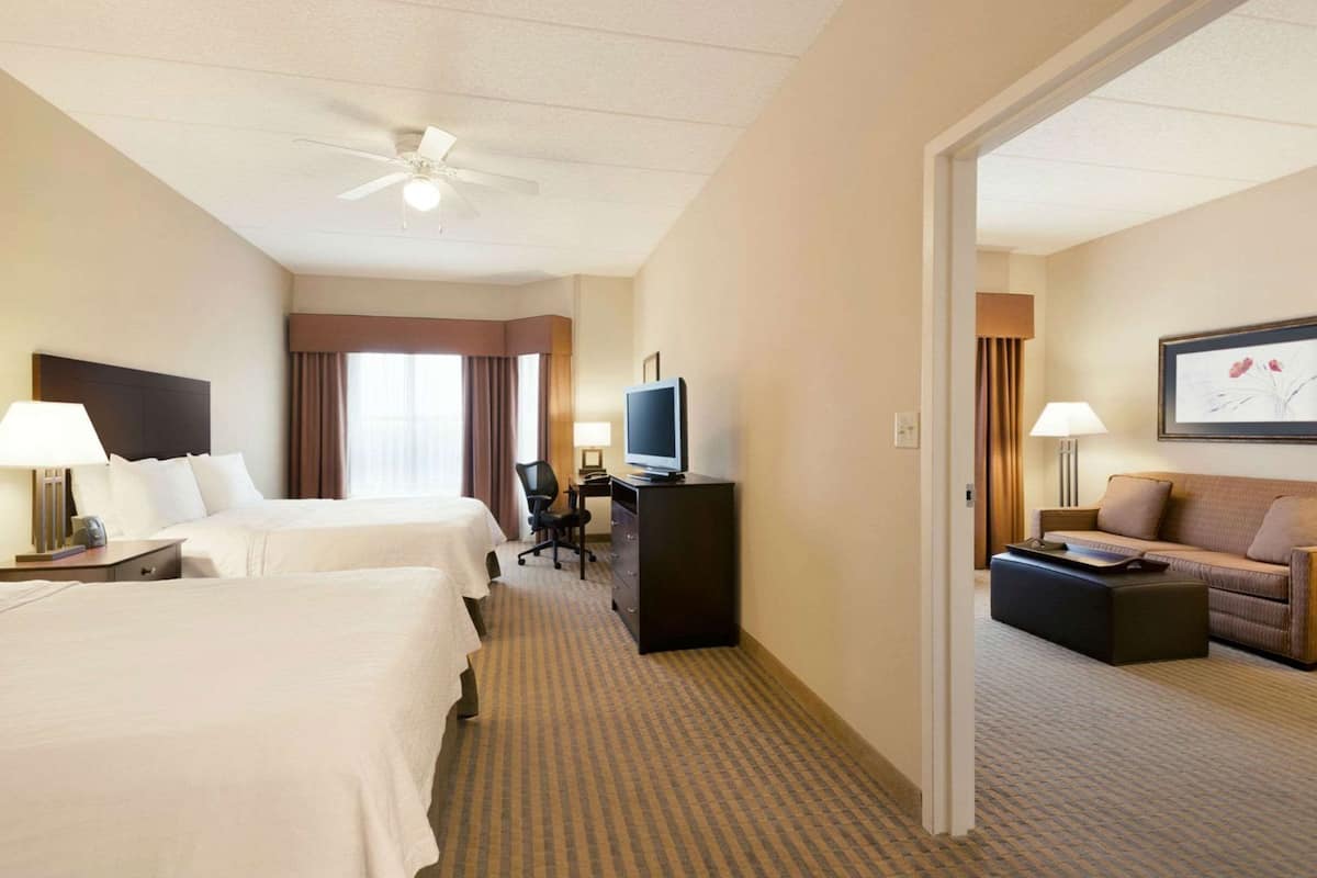 A hotel room with two beds, a desk with a television, a sofa, and a ceiling fan.