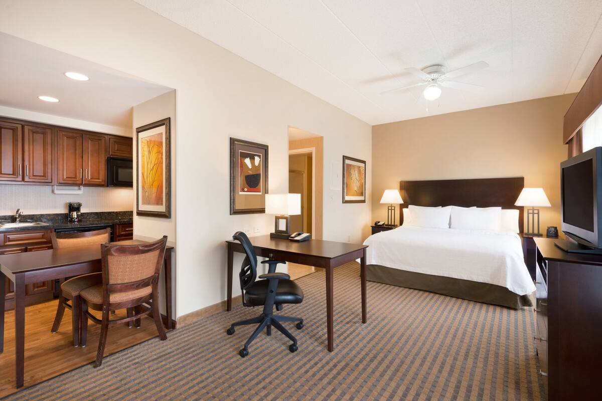 A hotel room with a bed, desk, chair, kitchenette, and ceiling fan.
