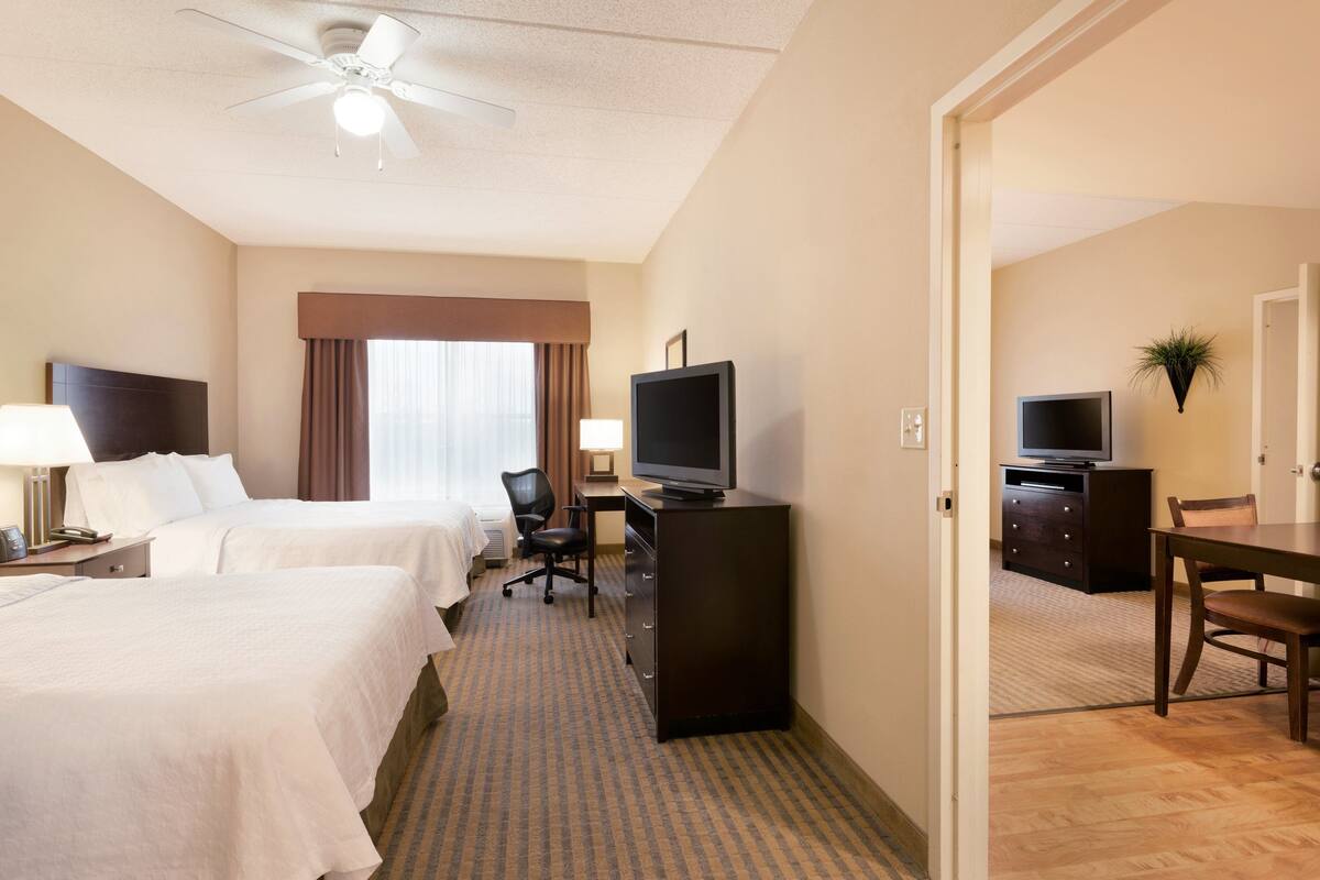 A hotel room with two beds, a desk with a TV, a chair, a desk lamp, and a ceiling fan.