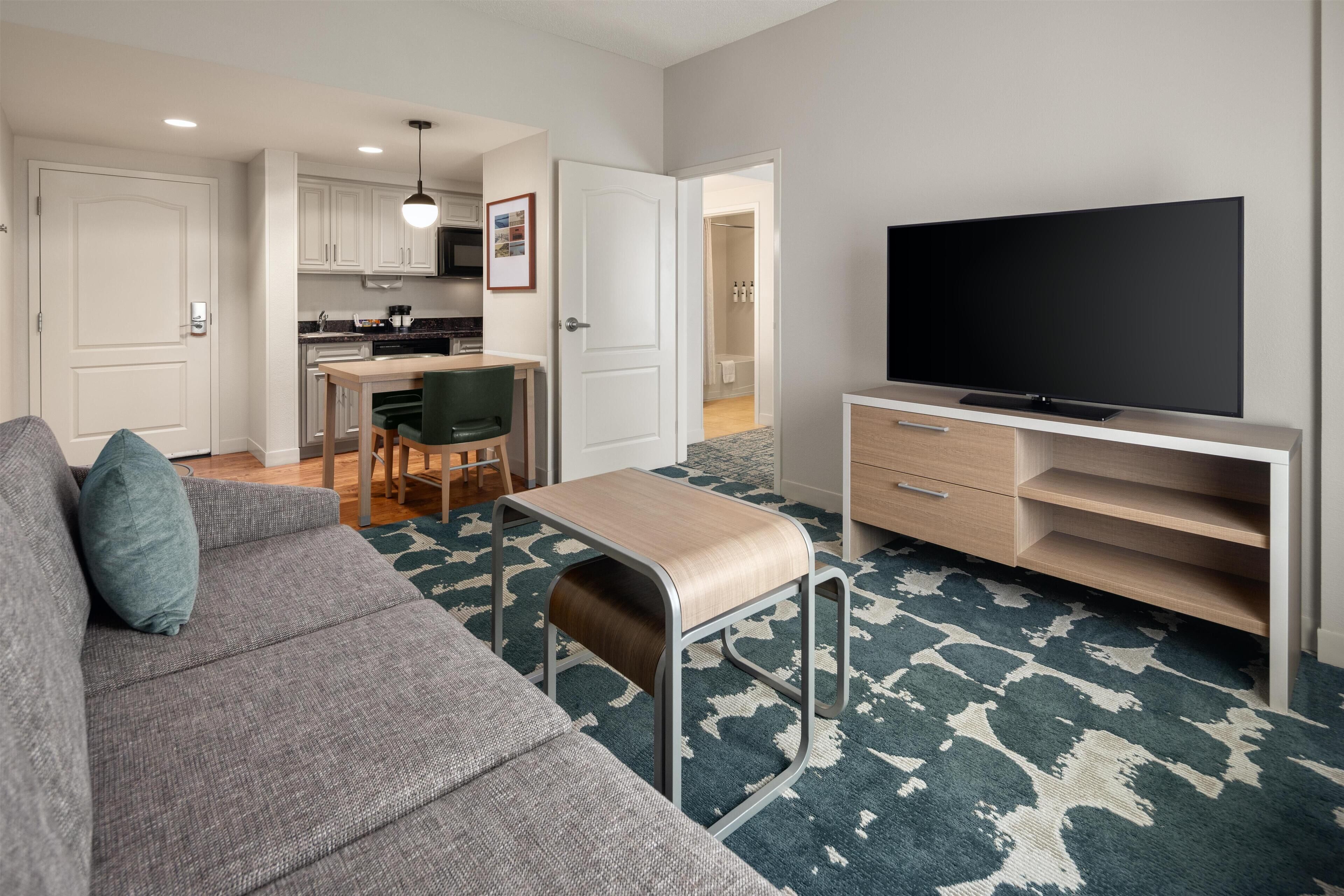 A modern living room with a sofa, a television on a stand, and a kitchen in the background.