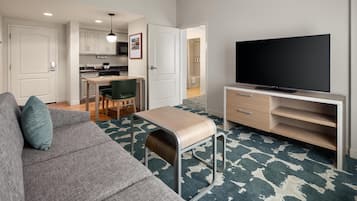 A modern living room with a sofa, a television on a stand, and a kitchen in the background.