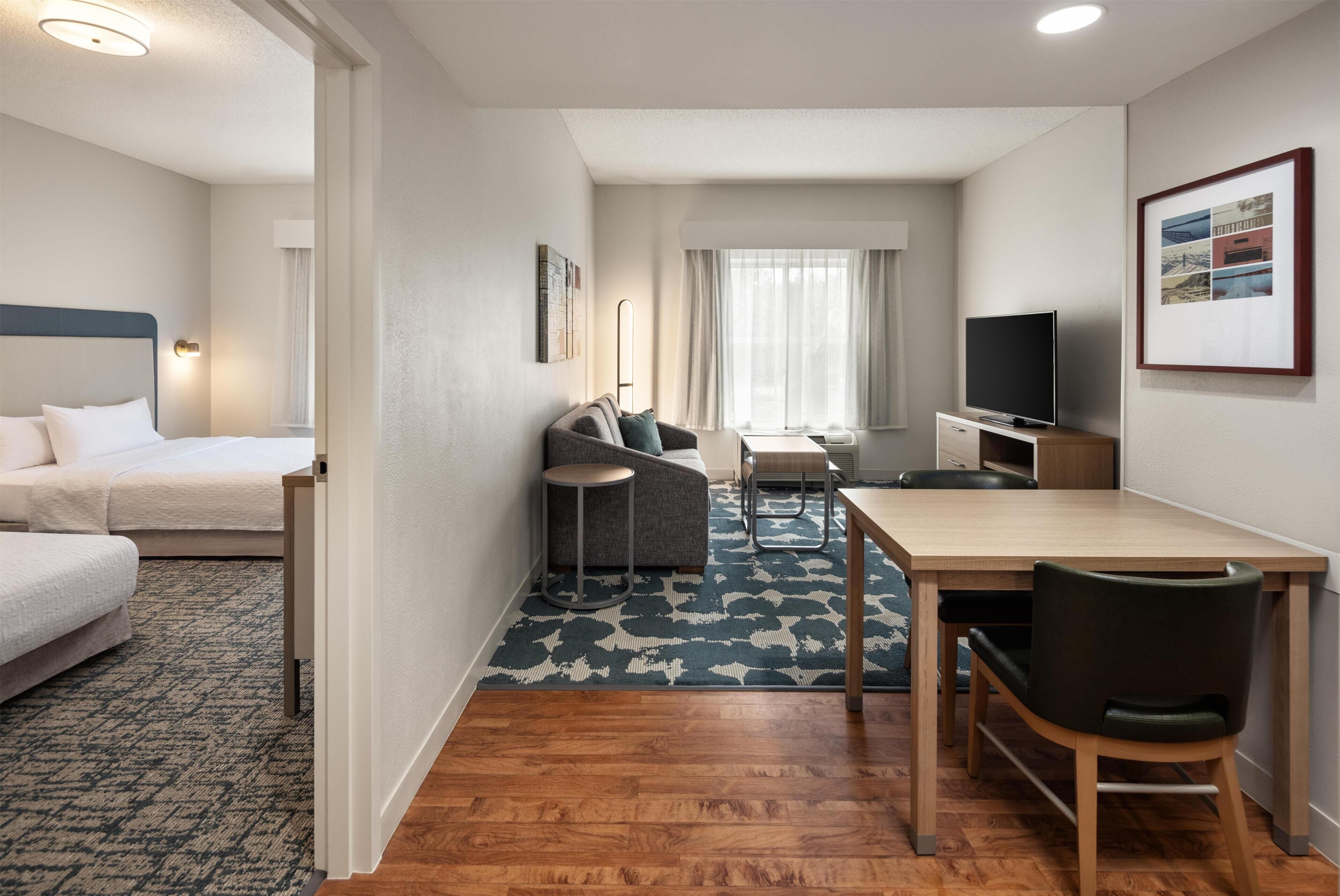 Habitación de hotel con cama, sofá, mesa de comedor con sillas, televisor y un amplio ventanal con cortinas.