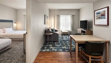 Habitación de hotel con cama, sofá, mesa de comedor con sillas, televisor y un amplio ventanal con cortinas.