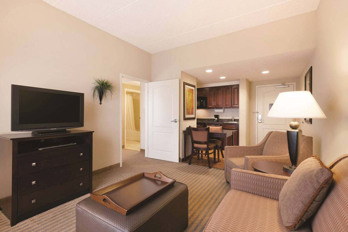 A hotel room with a flat-screen TV, a sofa, a coffee table, a dining area with chairs, and a kitchen area.