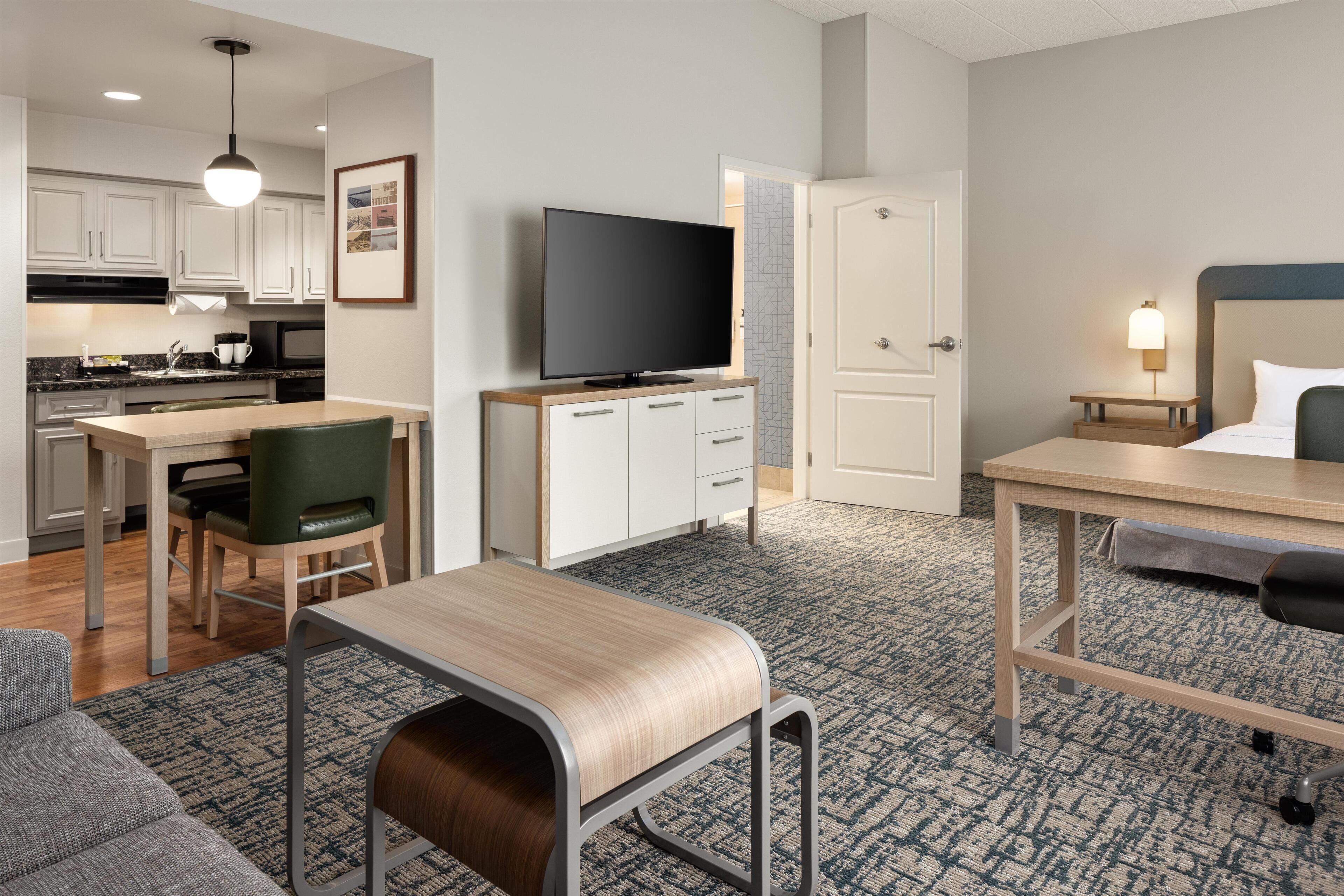Una habitación de hotel compacta con kitchenette, televisor de pantalla plana, cama con cabecera, pequeño escritorio y zona de estar con sillón y reposapiés.