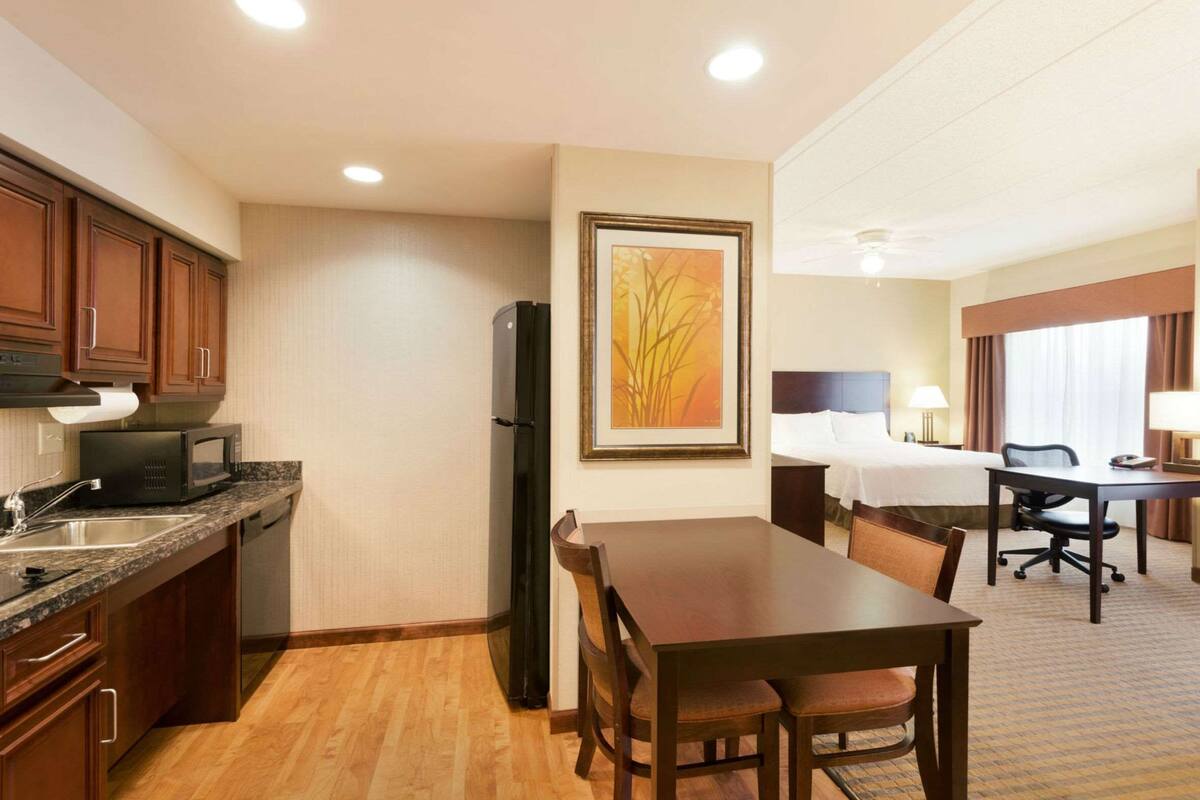 A hotel room with a kitchenette, a dining area, a bed, and a large window.