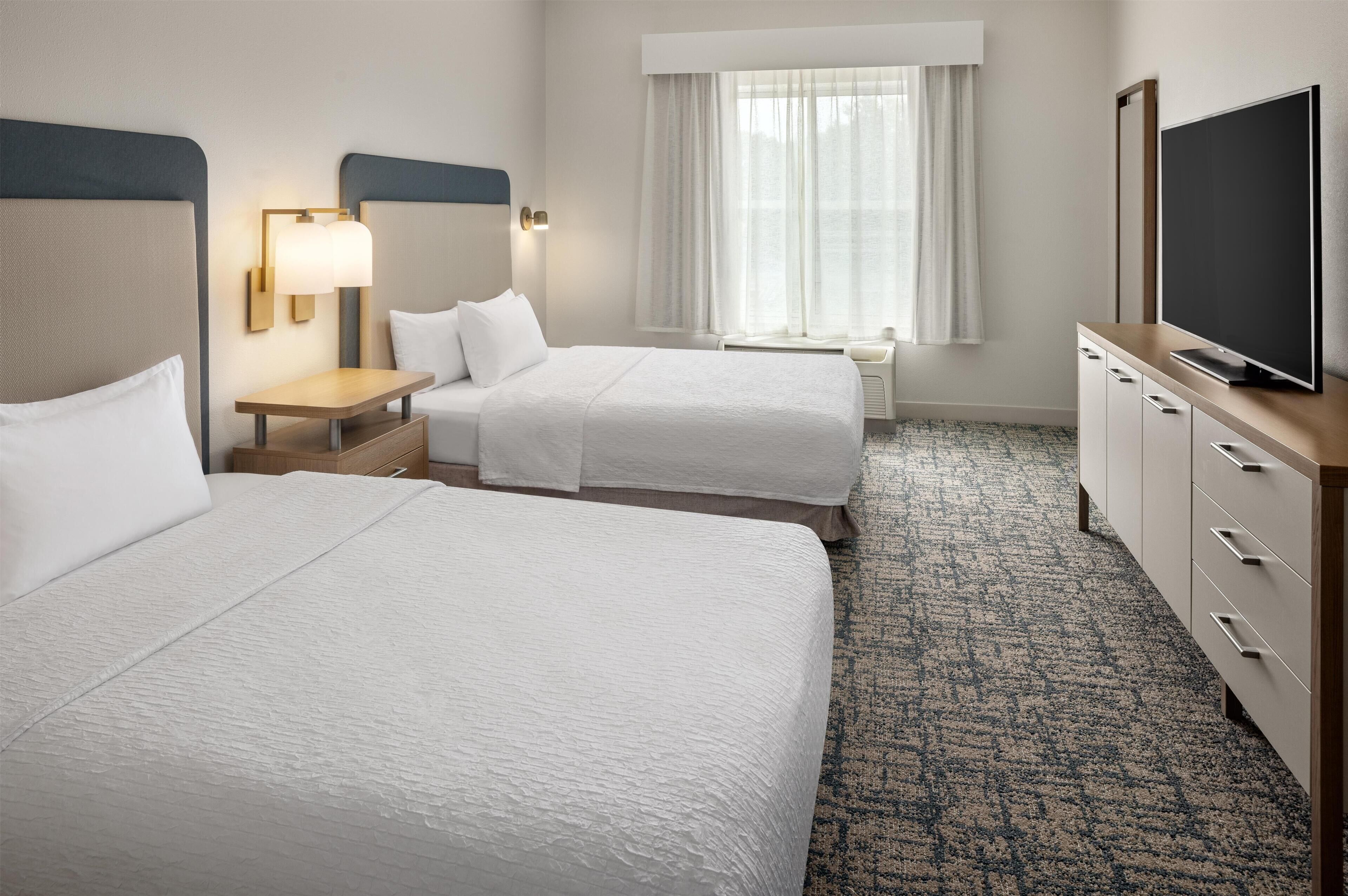 A hotel room with two beds, a desk, a TV, and a window with curtains.