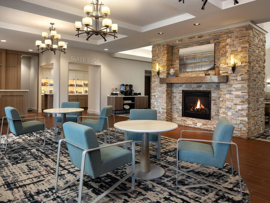 A hotel lobby with a stone fireplace, seating area with round tables and chairs, a kitchenette, and a 'Suite Shop' sign.