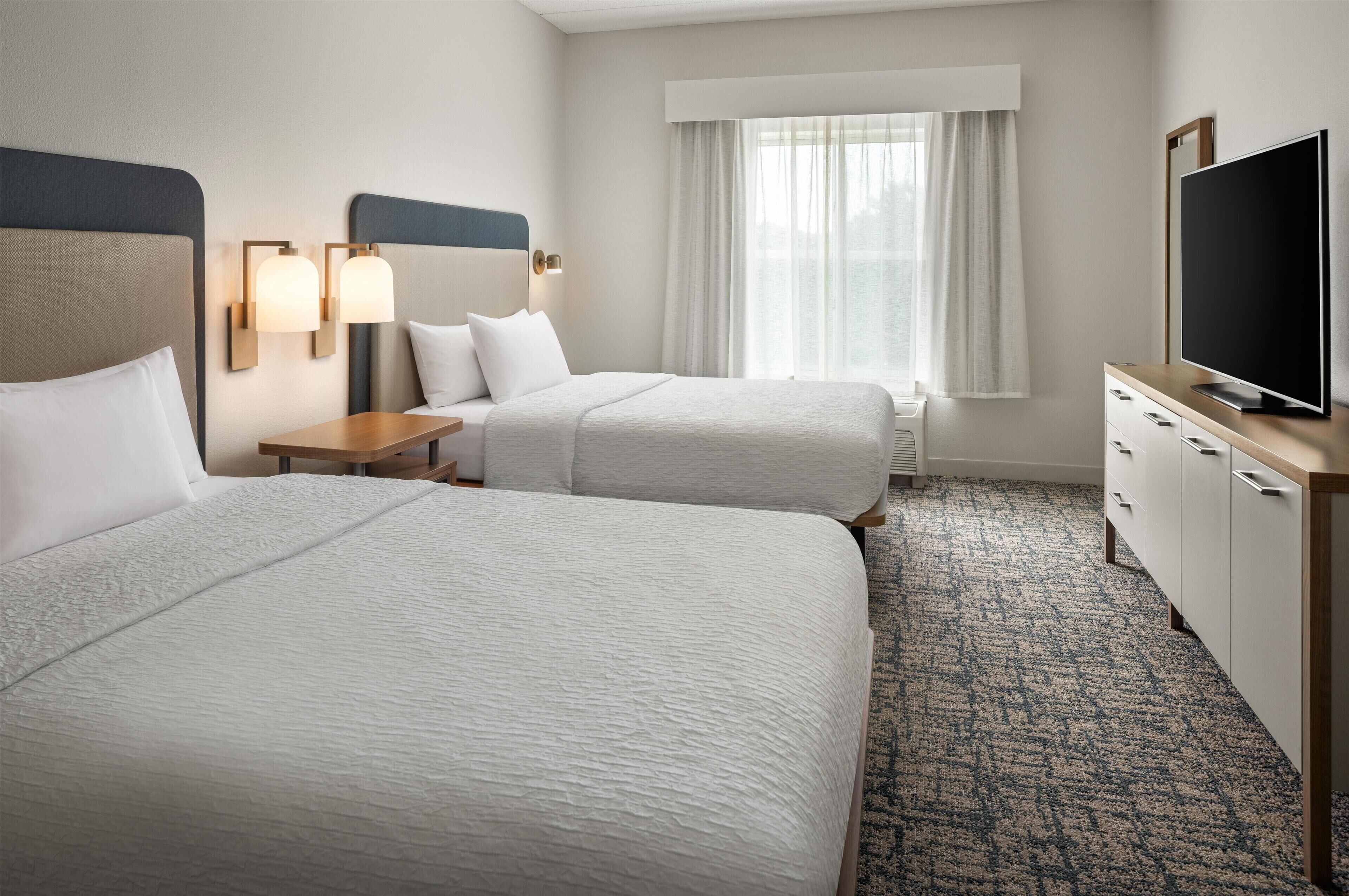 A hotel room with two beds, a television, a desk, and a window with curtains.