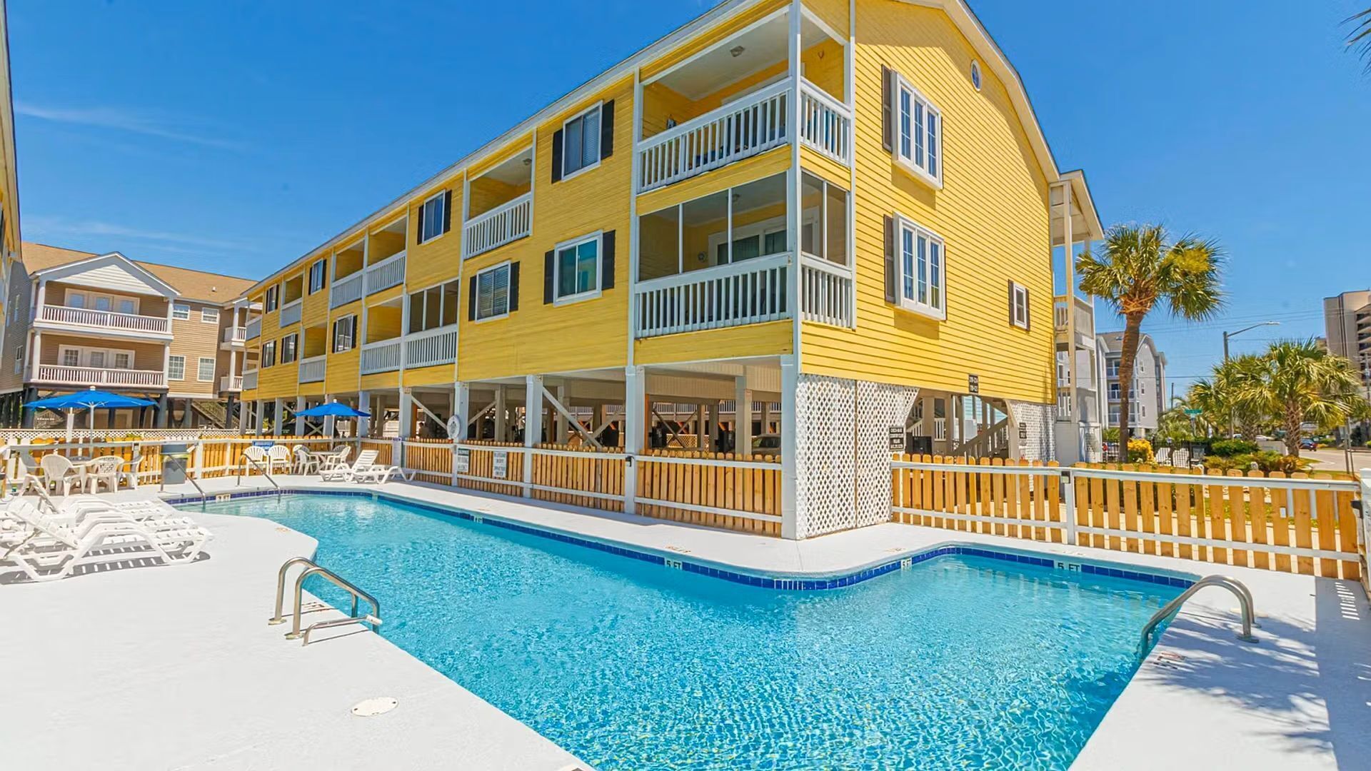 A yellow multi-story hotel with a pool area, including lounge chairs and umbrellas.
