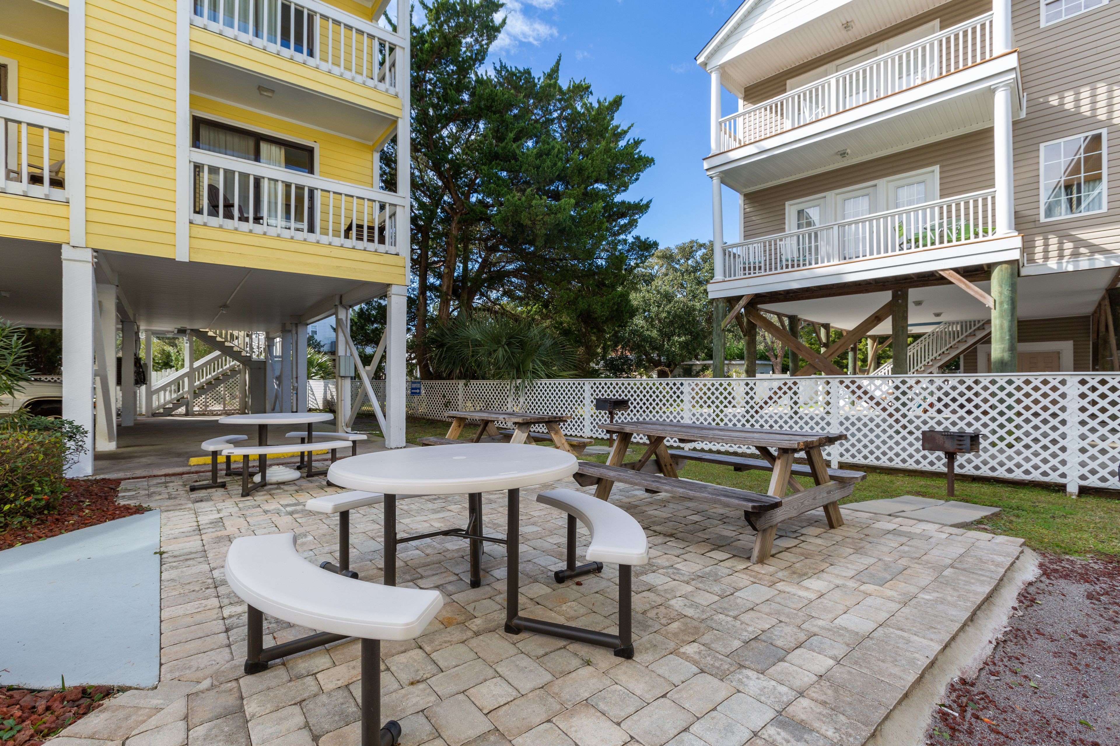 A patio area with picnic tables and benches, surrounded by a white fence and trees.