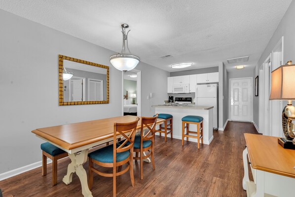A dining area with a wooden table and chairs, a kitchen in the background, and a hallway leading to a door.