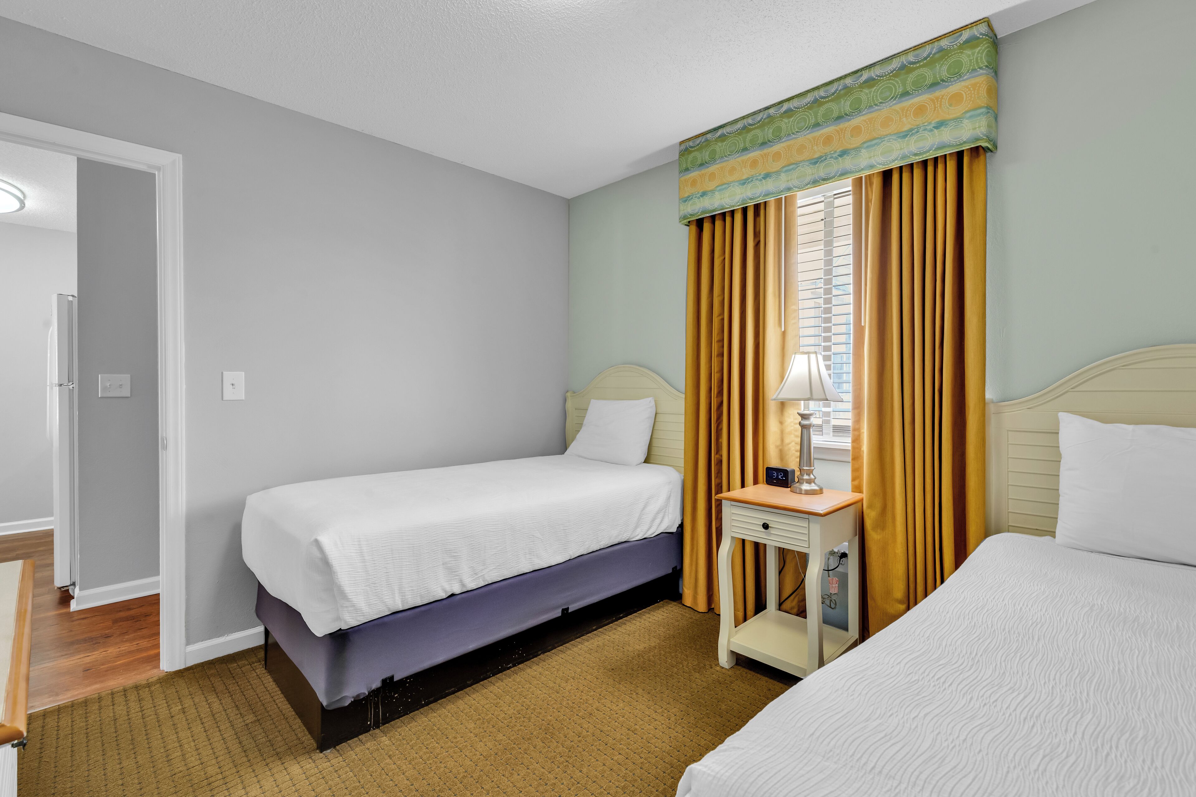 A hotel room with two beds, a bedside table with a lamp, and a window with curtains.