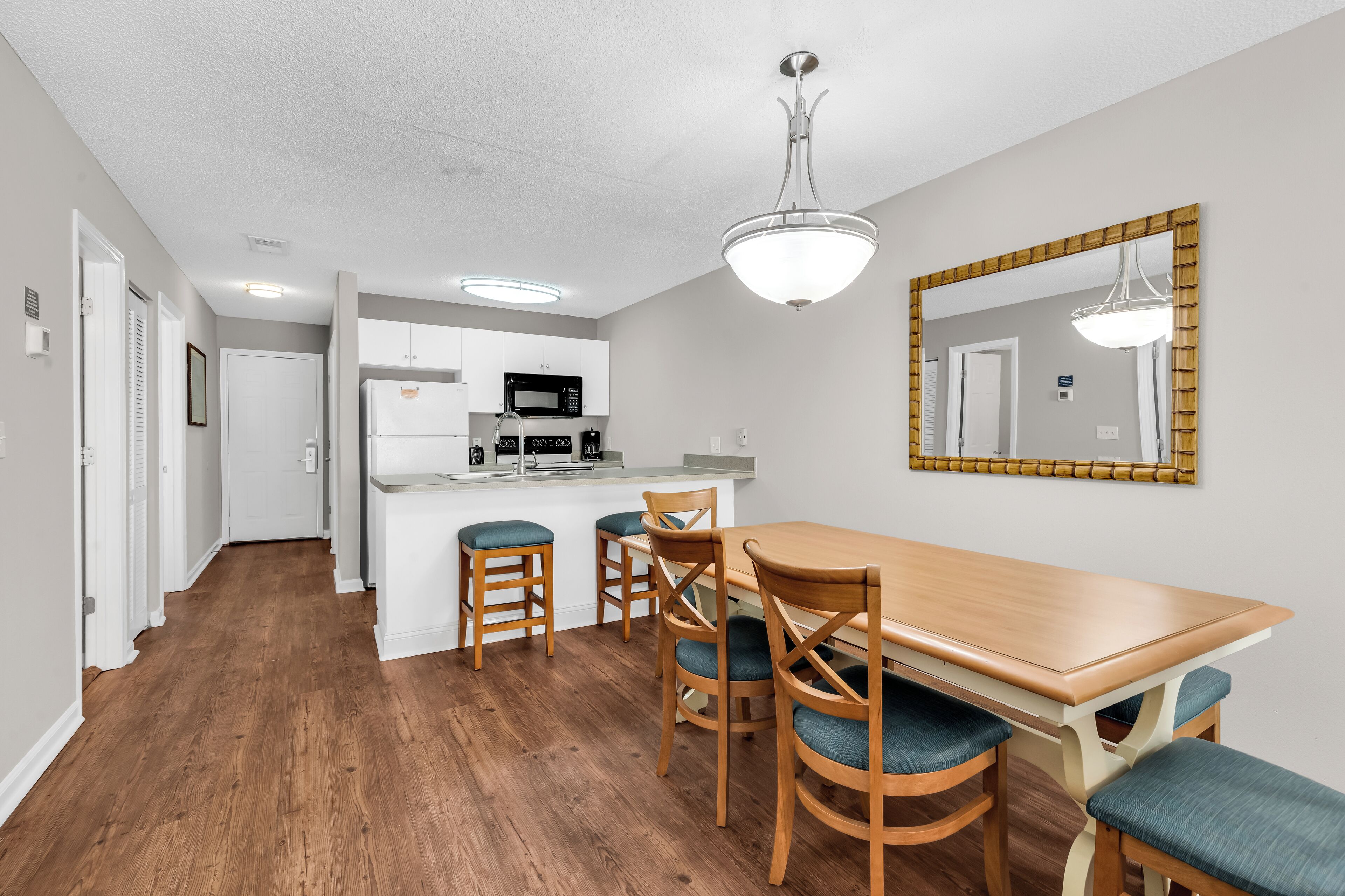 A modern kitchen with white cabinets, a microwave, and a sink. There is a wooden dining table with four chairs and a large mirror on the wall.
