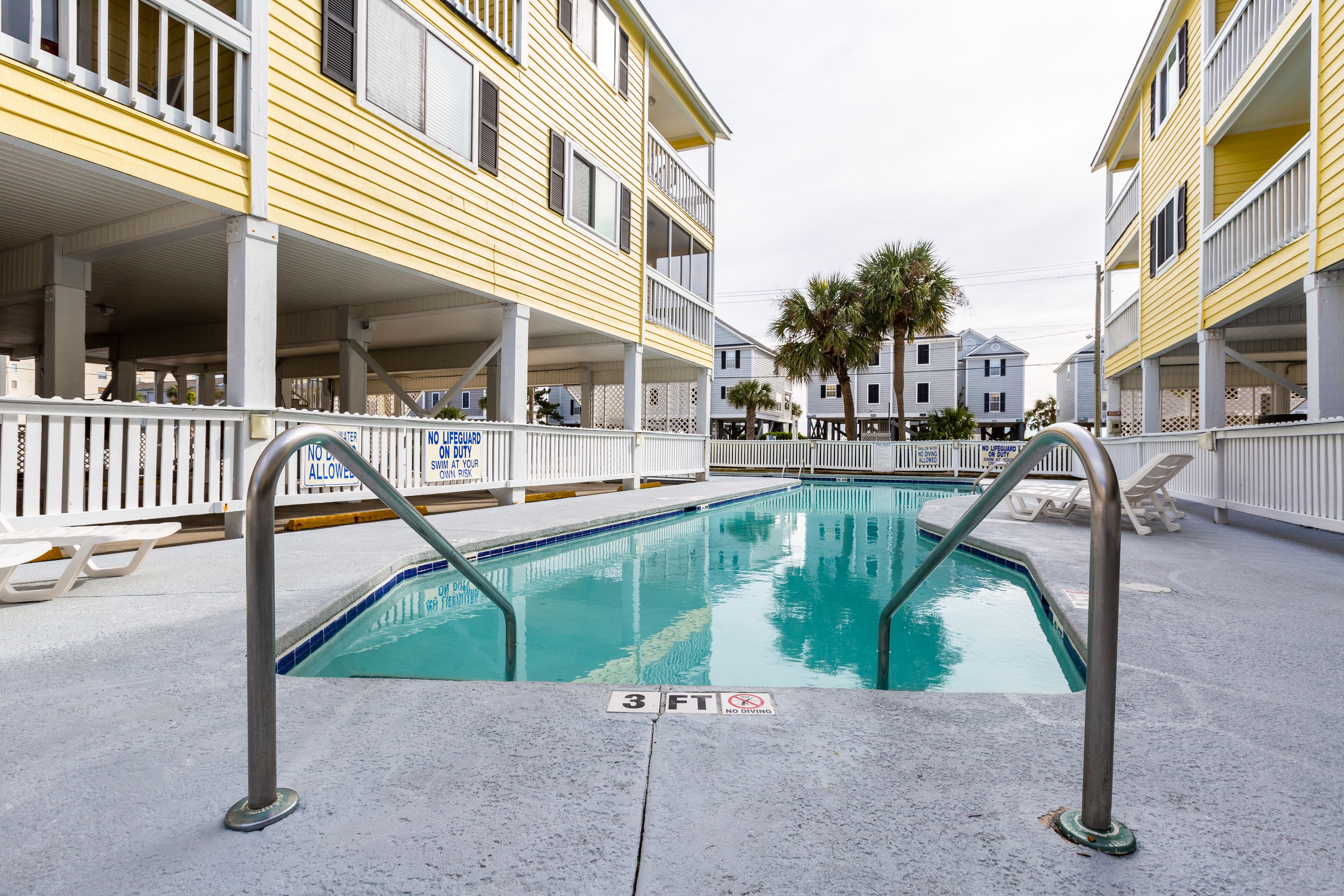 Une piscine dont la profondeur maximale est de 3 pieds, entourée d’une clôture blanche et bordée d’un bâtiment doté de balcons.