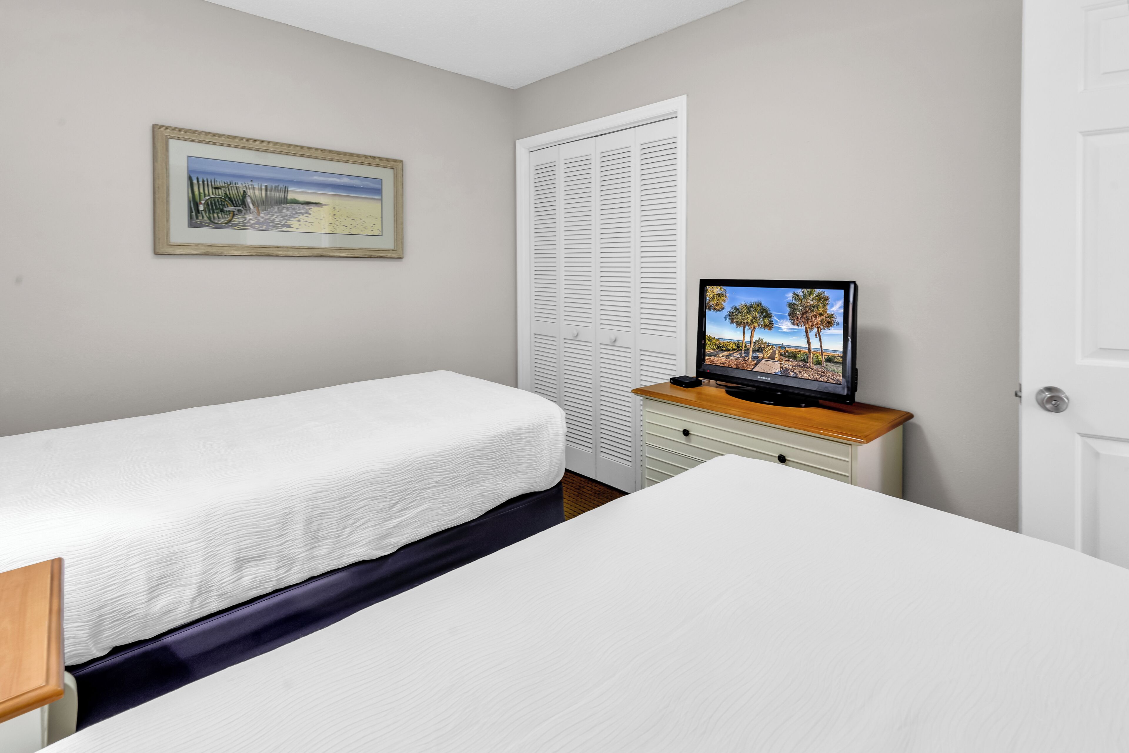 Une chambre avec deux lits, un téléviseur posé sur une commode en bois, une porte blanche et un tableau encadré accroché au mur.