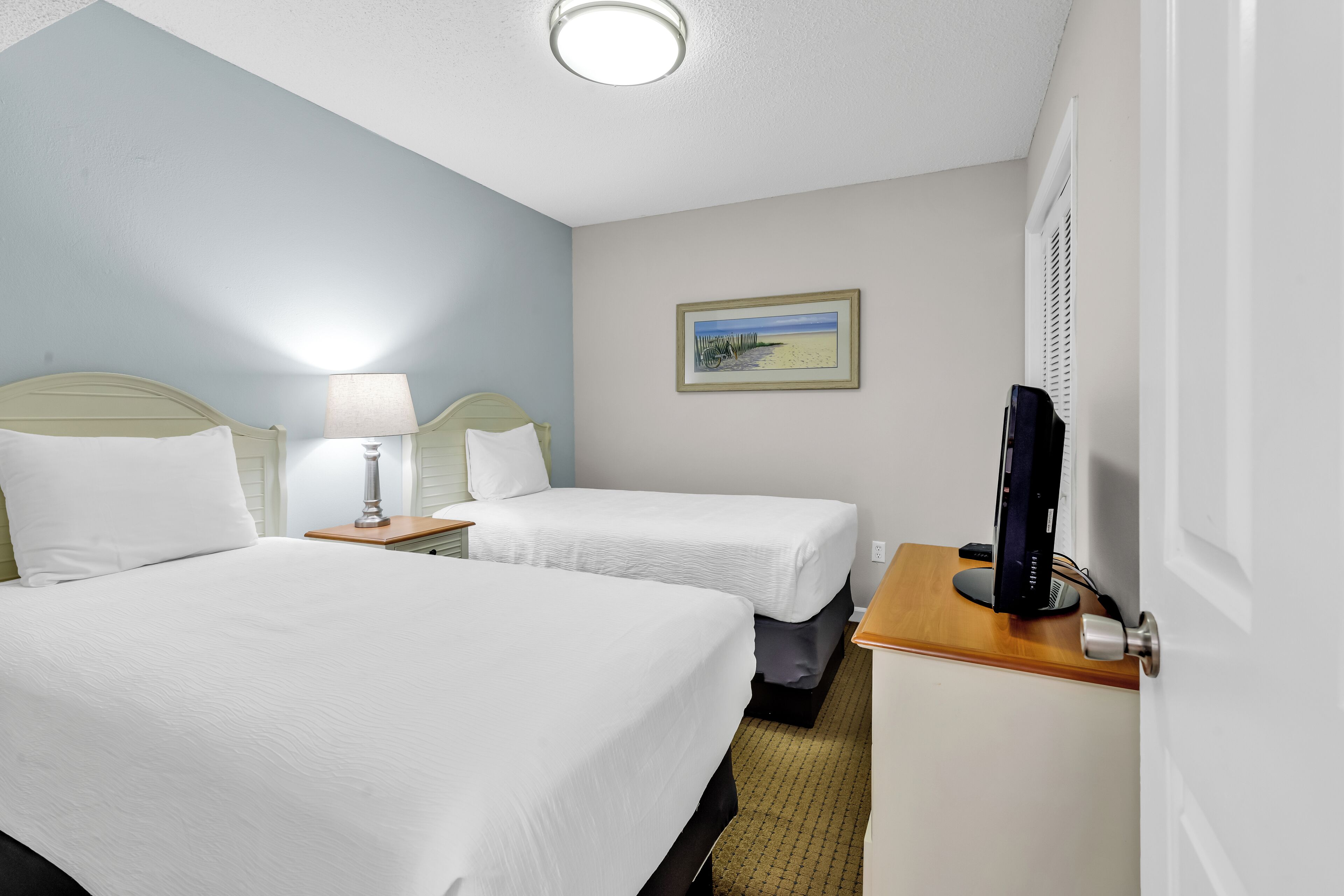 A hotel room with two beds, a desk, a computer, and a painting on the wall.
