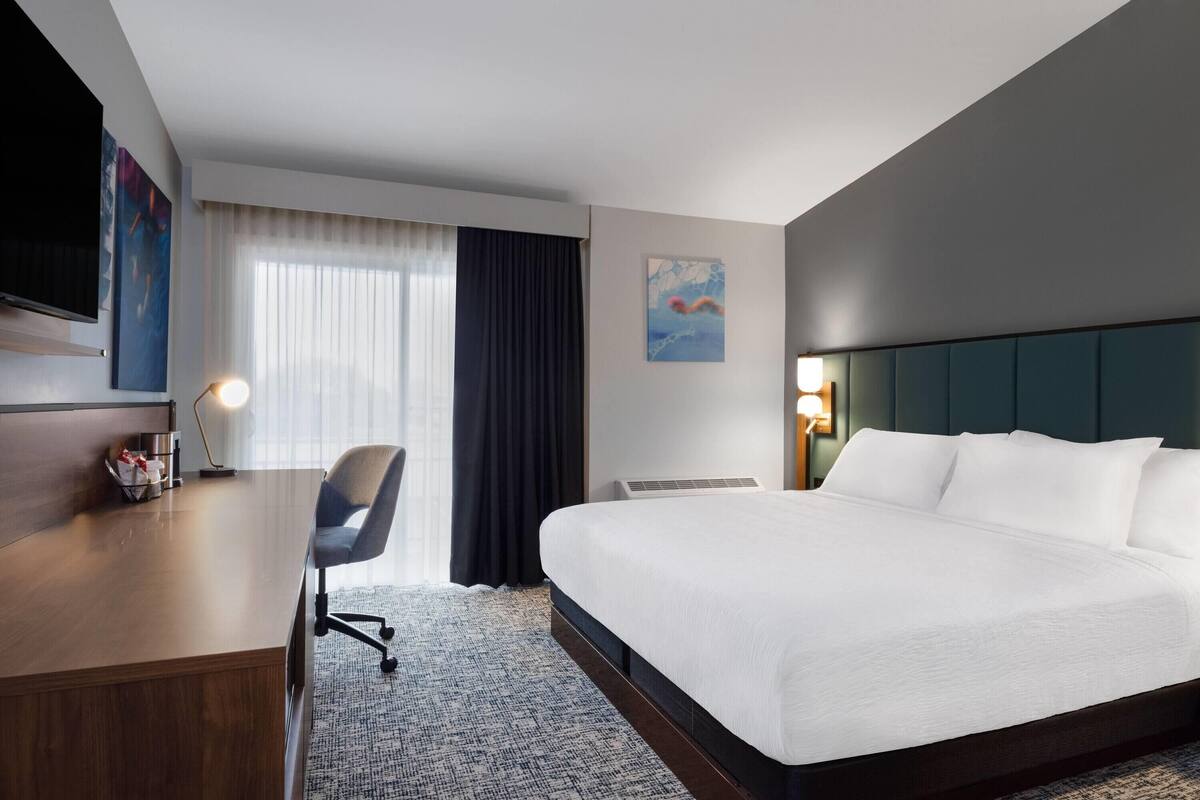 A hotel room with a large bed, a desk, a chair, a TV, and a painting on the wall.