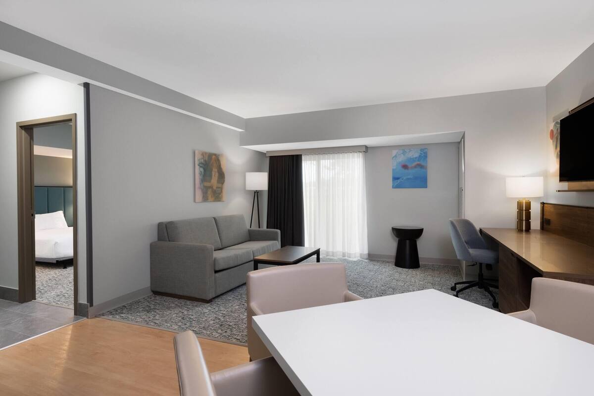 A hotel room with a dining area, a sofa, a TV, and a window with curtains.