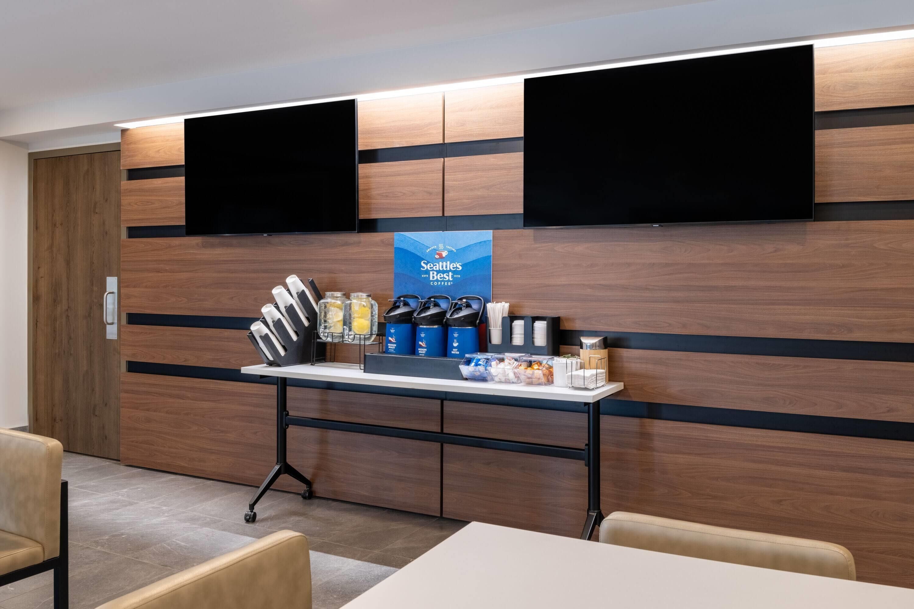 Zona d'entrada d'un hotel modern amb un panell de fusta a la paret, dos televisors penjats, una recepció amb serveis i una zona de seients.