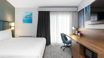Ein Hotelzimmer mit einem großen Bett, einem Schreibtisch mit Stuhl, einem Fernseher und einem Fenster mit Vorhängen.