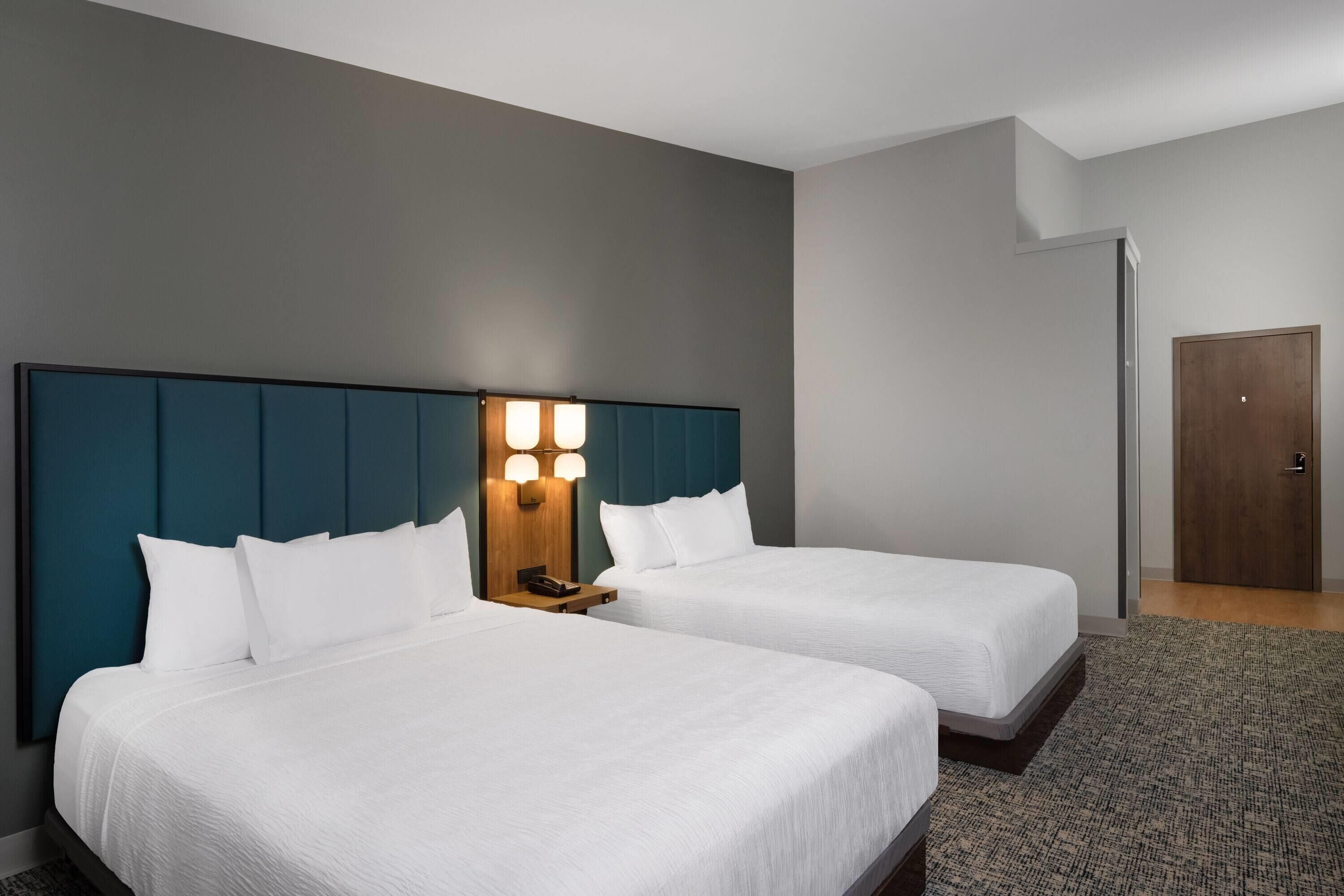 Ein Hotelzimmer mit zwei Betten, einem mit integrierter Beleuchtung versehenen Kopfteil, einer Holztür und einem Teppichboden.