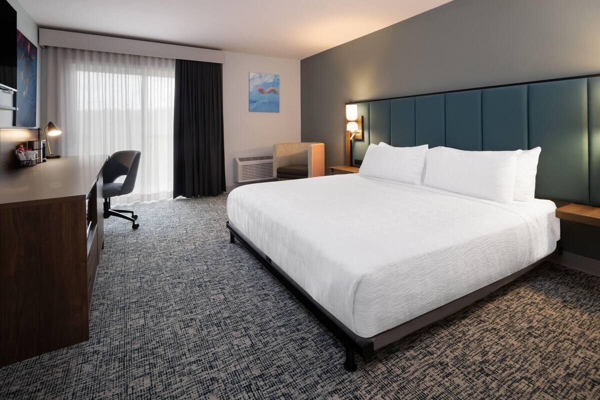 A hotel room with a large bed, a desk, a chair, a wall-mounted TV, and a painting.