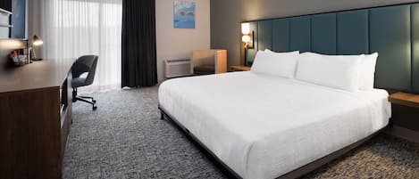 A hotel room with a large bed, a desk, a chair, a wall-mounted TV, and a painting.