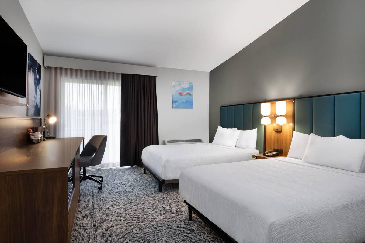 A hotel room with two beds, a desk, a chair, a flat-screen TV, and a painting on the wall.
