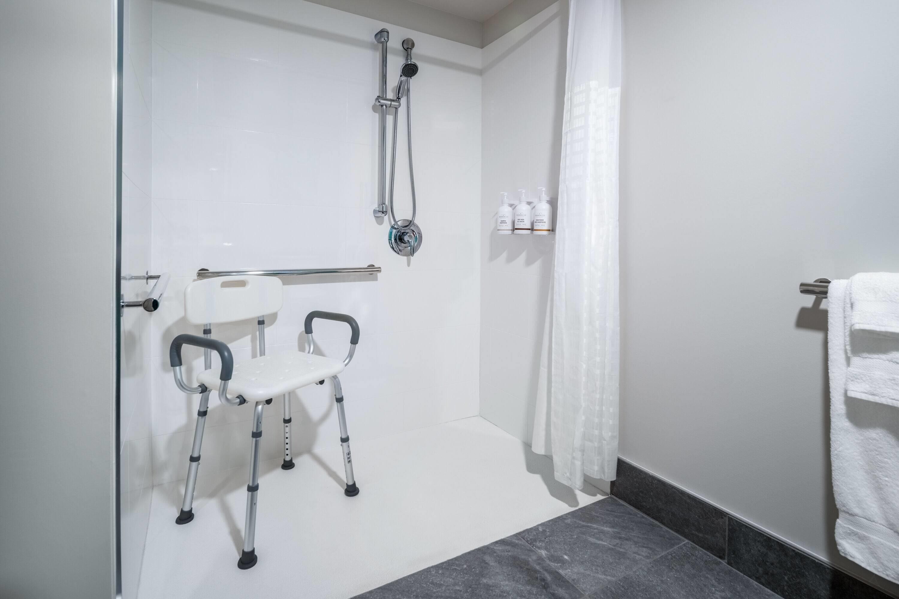 A bathroom with a shower, a handrail, a white chair, and a towel rack.