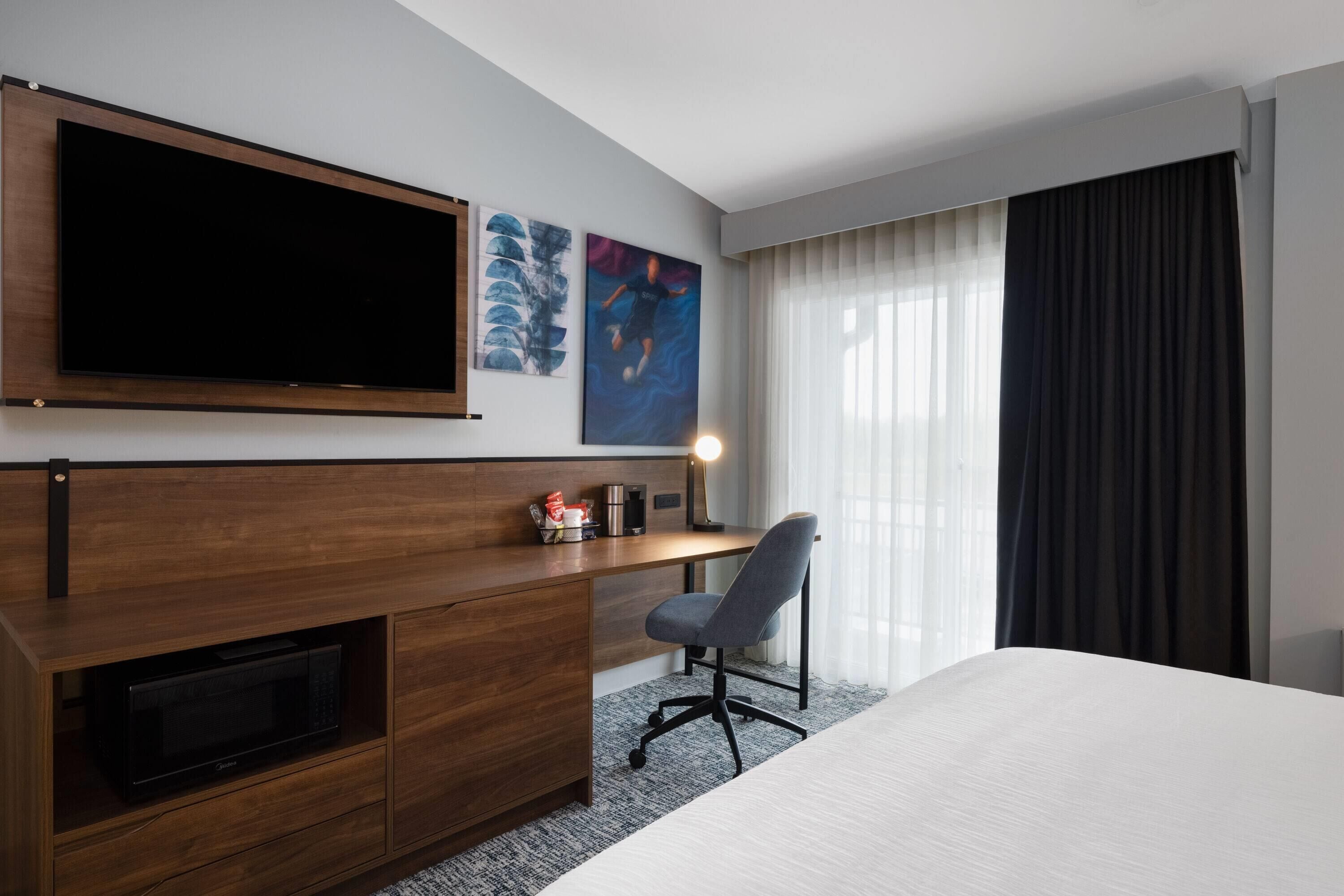 A hotel room with a bed, a desk with a chair, a flat-screen TV, a microwave, artwork on the wall, and a window with curtains.