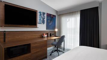 A hotel room with a bed, a desk with a chair, a flat-screen TV, a microwave, artwork on the wall, and a window with curtains.