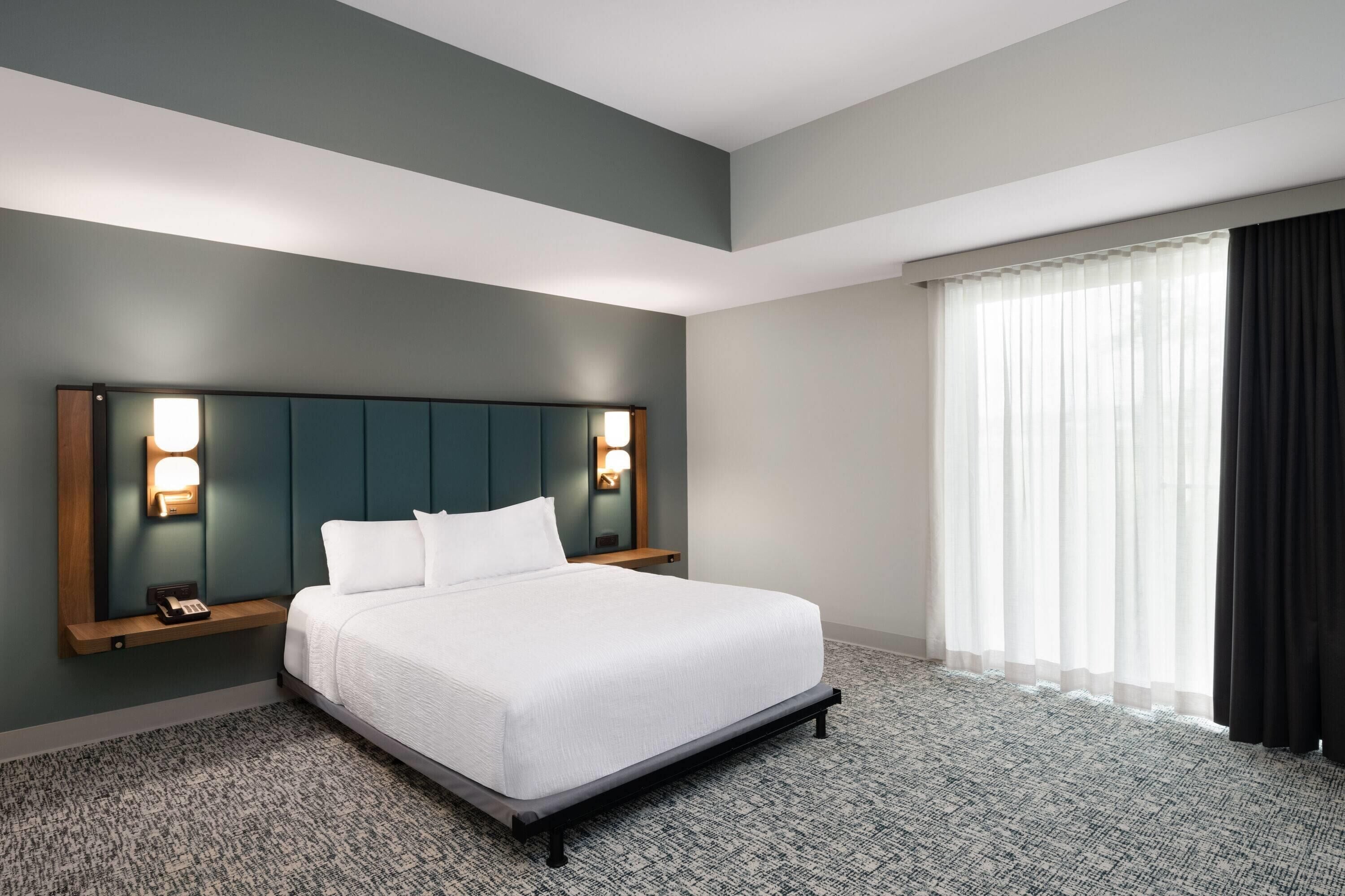 Une chambre d’hôtel avec un grand lit, une tête de lit dotée de lampes fixées au mur, une table de chevet avec un téléphone et une fenêtre ornée de rideaux légers.